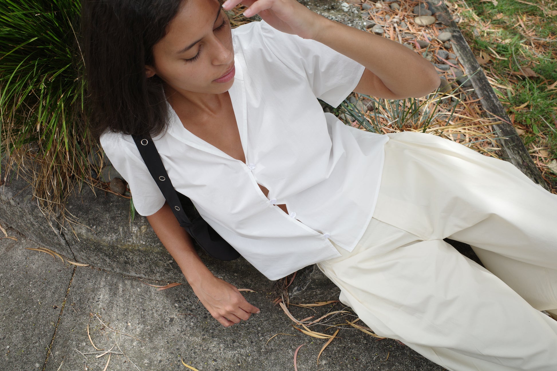 A woman with medium-length dark hair sits outdoors on a curb, wearing the Sails Top - White and loose cream pants. She looks down, touching her hair, surrounded by dried leaves and grass.