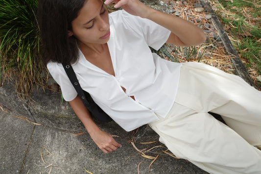 A woman with medium-length dark hair sits outdoors on a curb, wearing the Sails Top - White and loose cream pants. She looks down, touching her hair, surrounded by dried leaves and grass.
