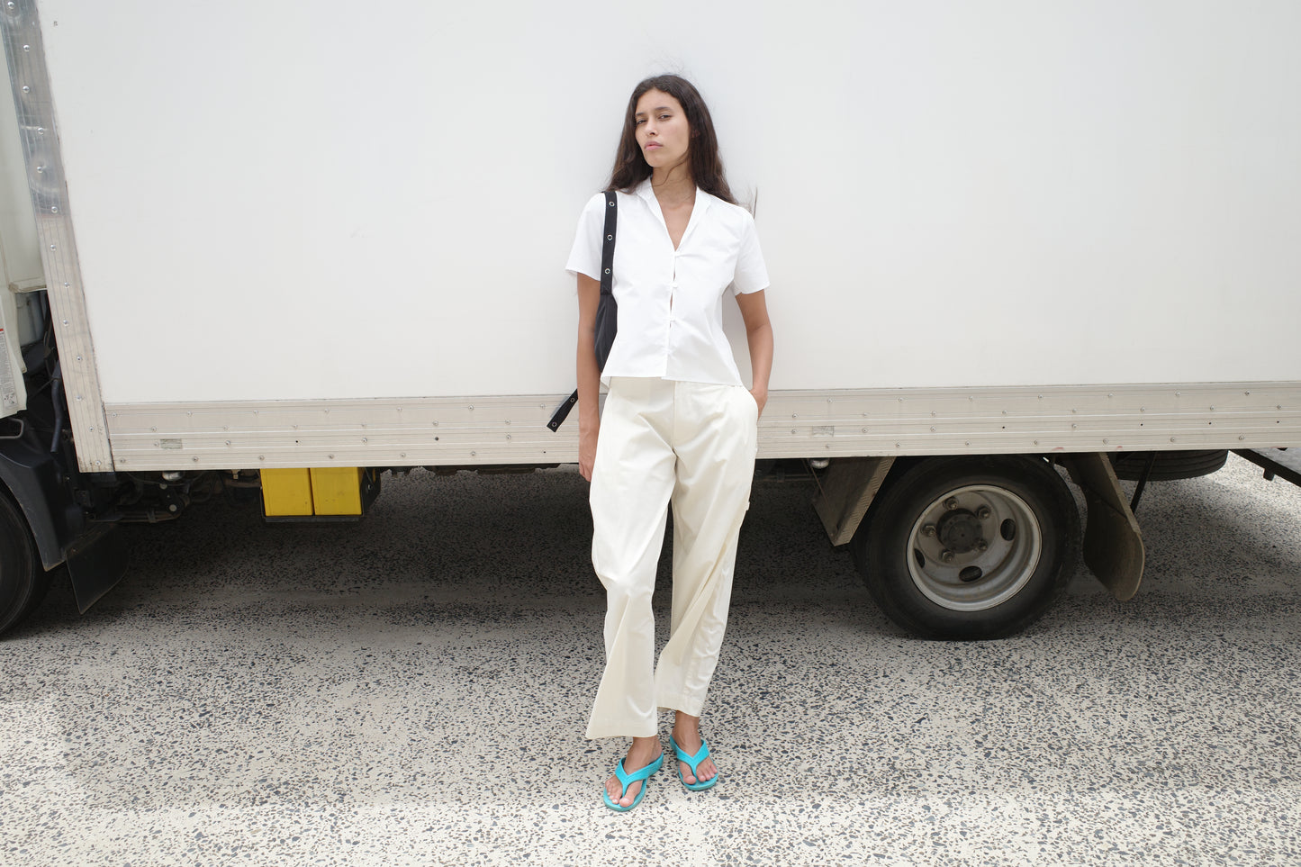 A woman stands in front of a white truck, wearing the Sails Top - White, light pants, teal flip-flops, and a black shoulder bag. She has her hands in her pockets and looks to the side.