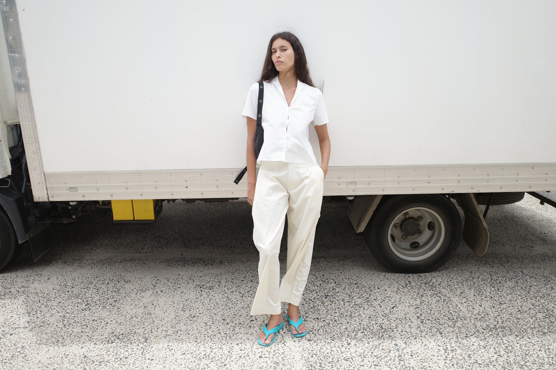 A woman stands in front of a white truck, wearing the Sails Top - White, light pants, teal flip-flops, and a black shoulder bag. She has her hands in her pockets and looks to the side.