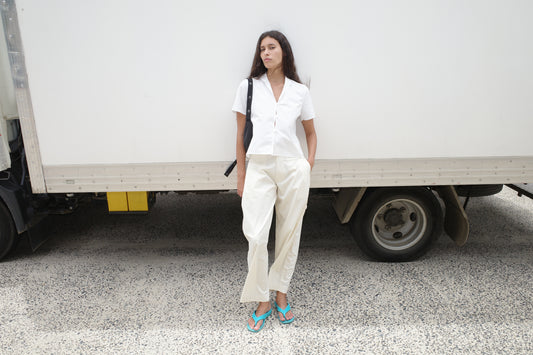 A woman stands in front of a white truck, wearing the Sails Top - White, light pants, teal flip-flops, and a black shoulder bag. She has her hands in her pockets and looks to the side.