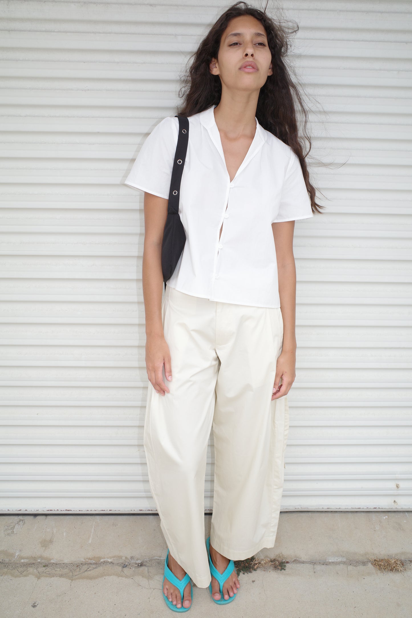 A woman with long, loose hair and a relaxed expression stands before a white metal shutter, wearing an oversized Sails Top - White made from organic cotton, wide-legged cream pants, teal sandals, and carrying a black shoulder bag.