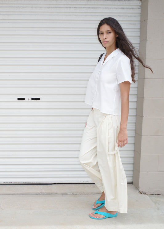 A woman with long brown hair stands in front of a white rolling door, wearing the Sails Top - White, loose cream pants, blue flip-flops, and carrying a black bag on her shoulder.