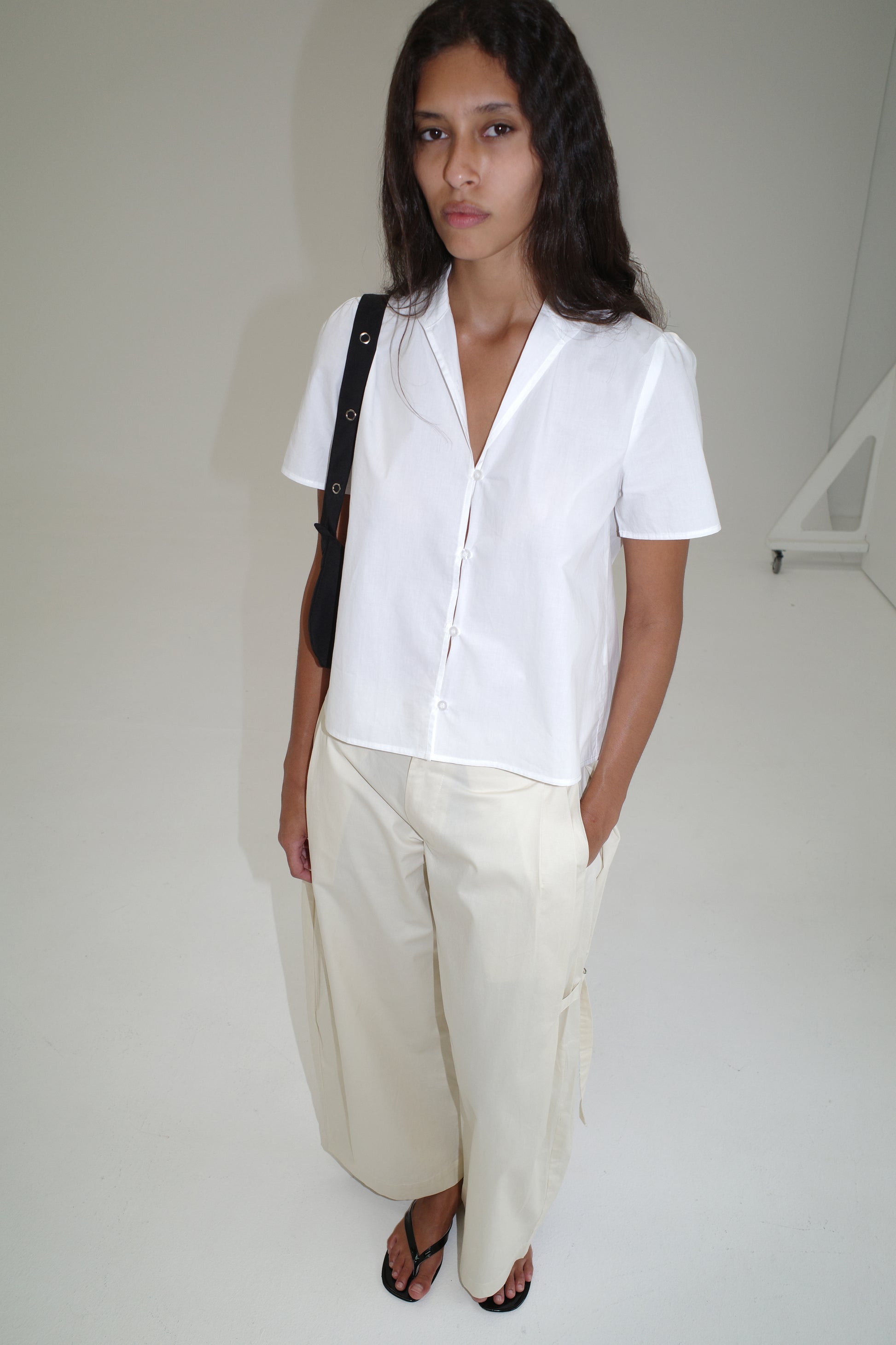 A person with long dark hair wears the Sails Top - White, wide cream pants, black sandals, and carries a black shoulder bag, standing against a plain white background.
