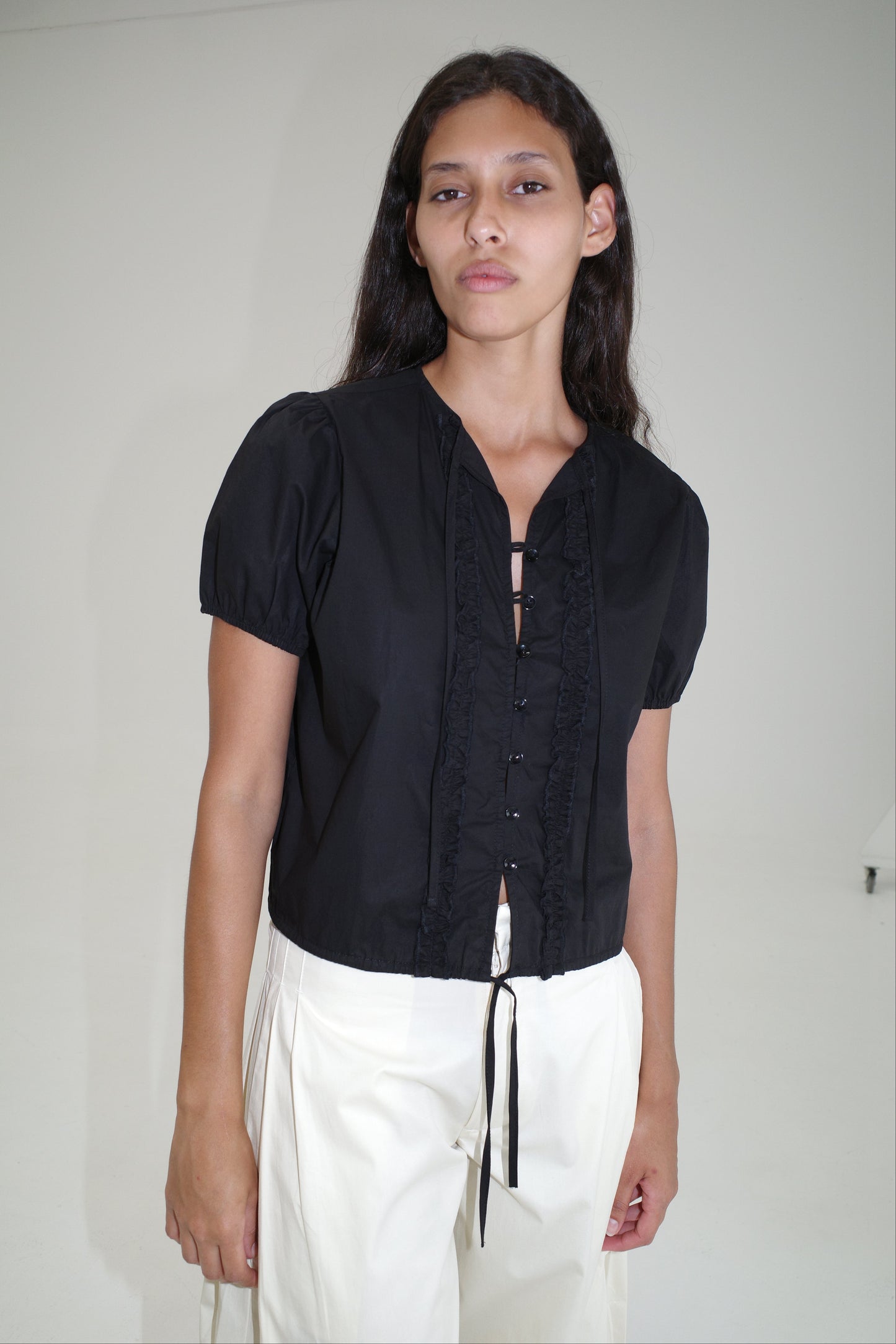 A woman with long dark hair wears the Double Frill Top - Black by Deiji Studios and white high-waisted pants, standing against a plain white background.