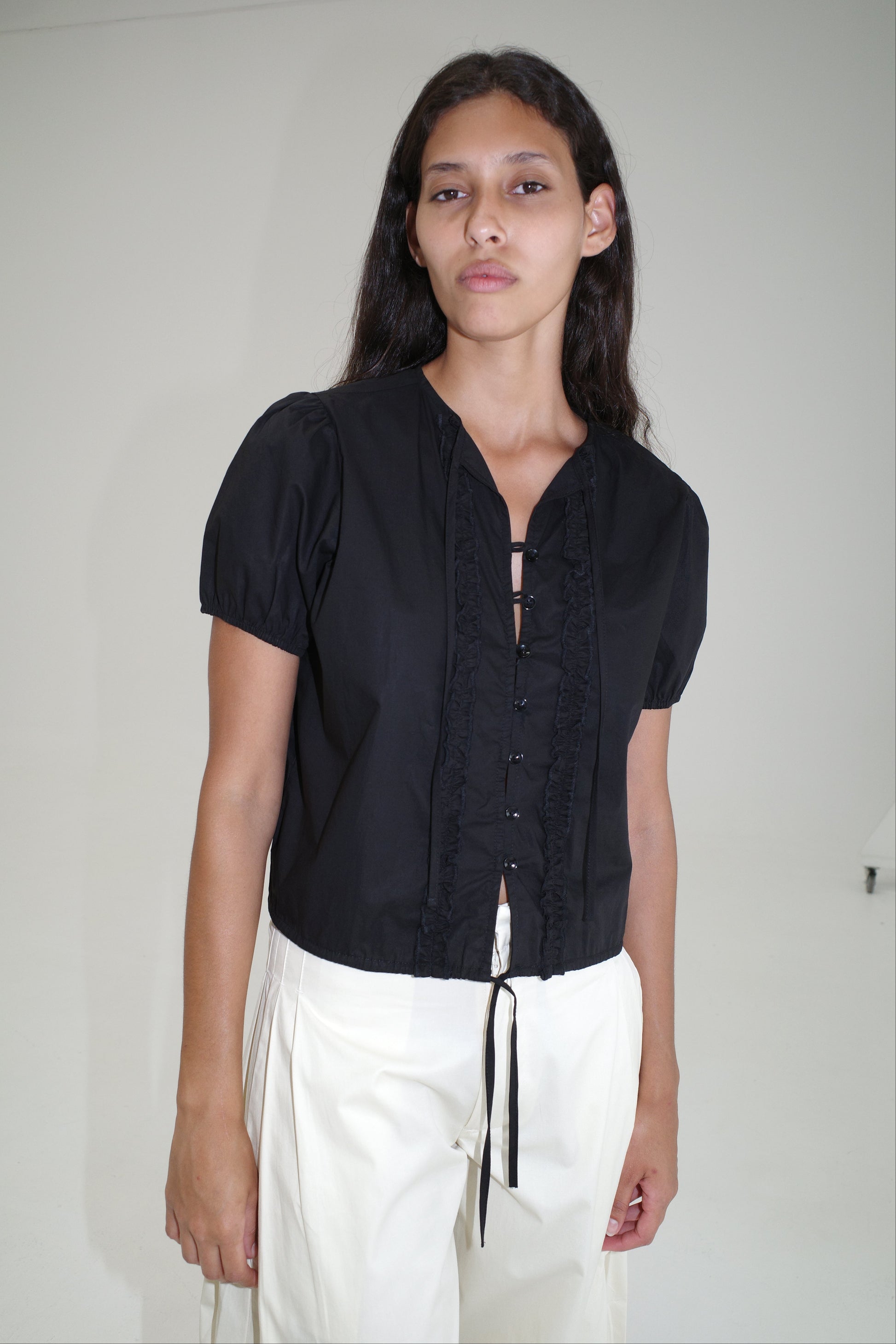 A woman with long dark hair wears the Double Frill Top - Black by Deiji Studios and white high-waisted pants, standing against a plain white background.