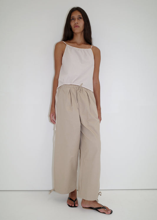 A woman with long, straight hair and a relaxed posture stands against a white background in the Drawcord Wide Leg Pant - Cream by Deiji Studios, paired with a sleeveless top and black flip-flops.