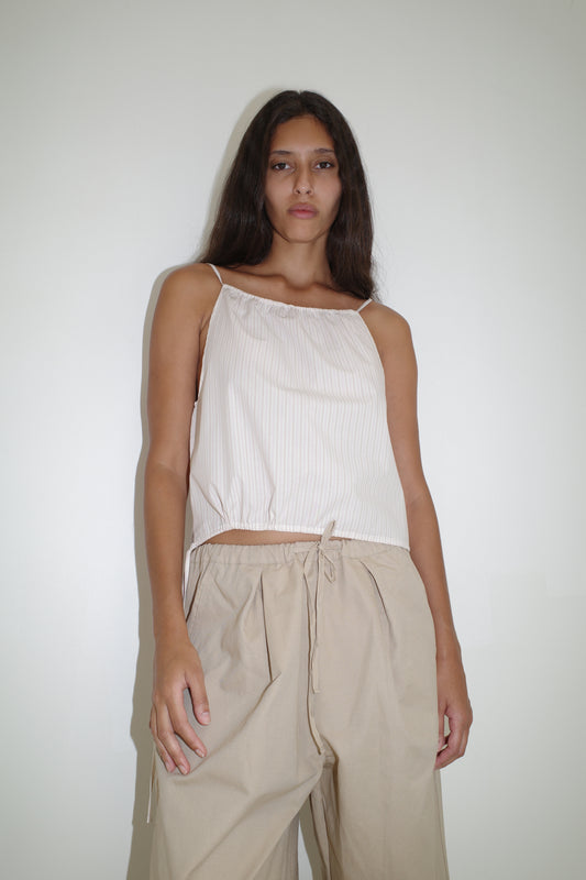 A person with long dark hair wears a sleeveless light top and the Drawcord Wide Leg Pant - Cream by Deiji Studios in organic cotton, standing against a plain white background and looking at the camera.
