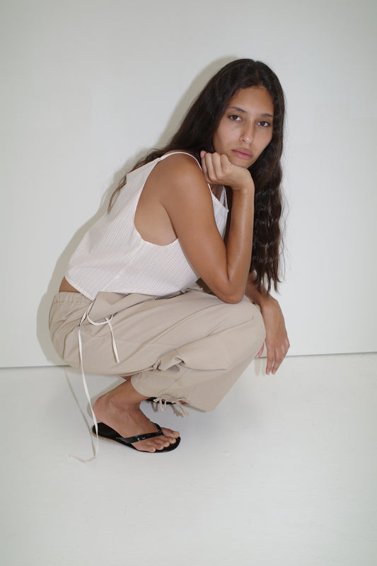 A person with long, wavy hair crouches by a white wall, wearing a Deiji Studios white sleeveless top and the Drawcord Wide Leg Pant in Cream, paired with black sandals, resting their chin on one hand and looking at the camera.