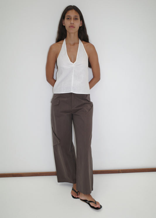 A woman with long dark hair stands against a white wall, arms behind her back, wearing a Deiji Studios Linen Pinched Halter Top in white, loose brown pants with pockets, and black flip-flops.