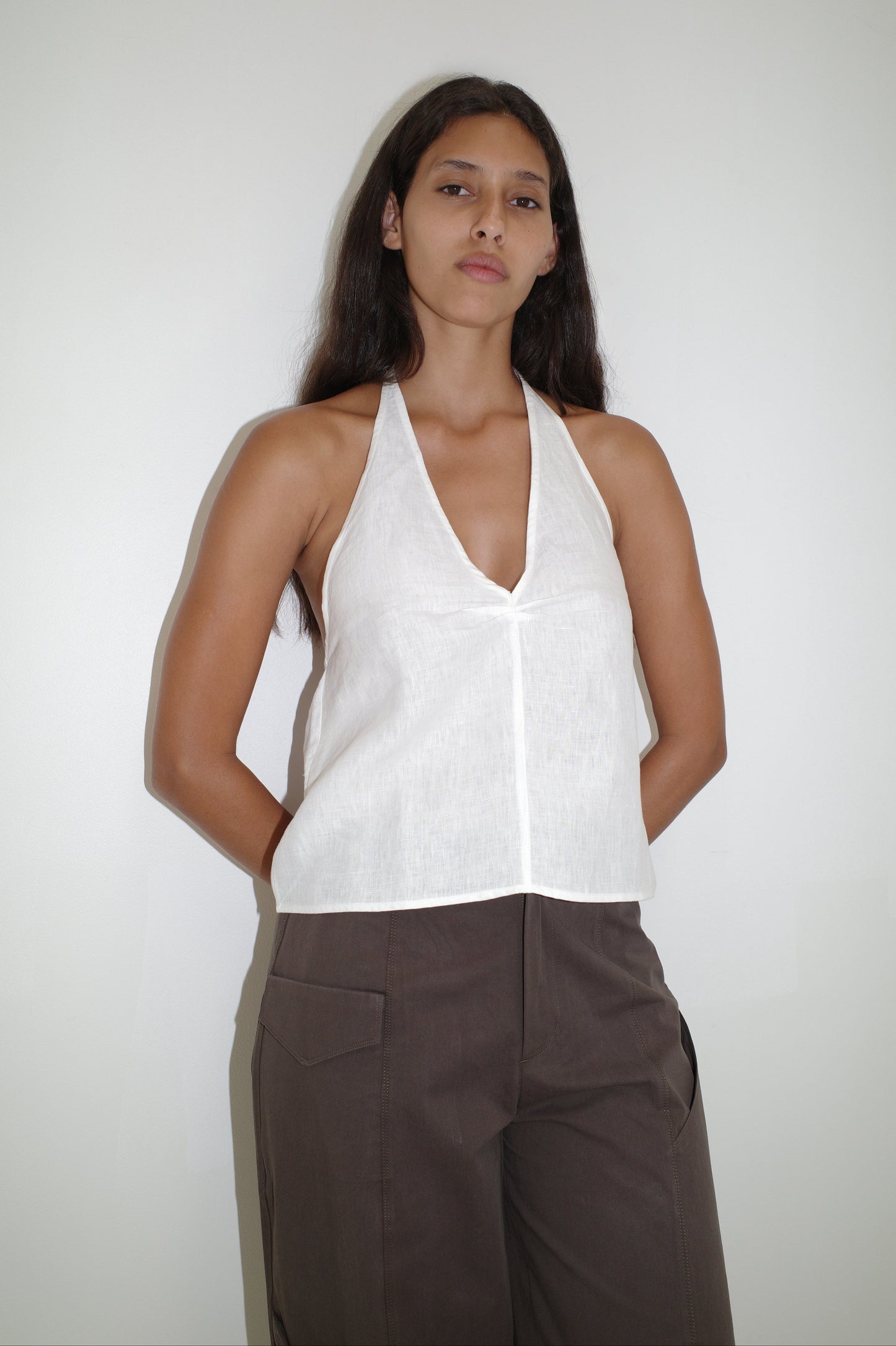A person with long dark hair stands against a plain white wall, wearing the Linen Pinched Halter Top - White and brown pants, hands behind their back and a neutral facial expression.