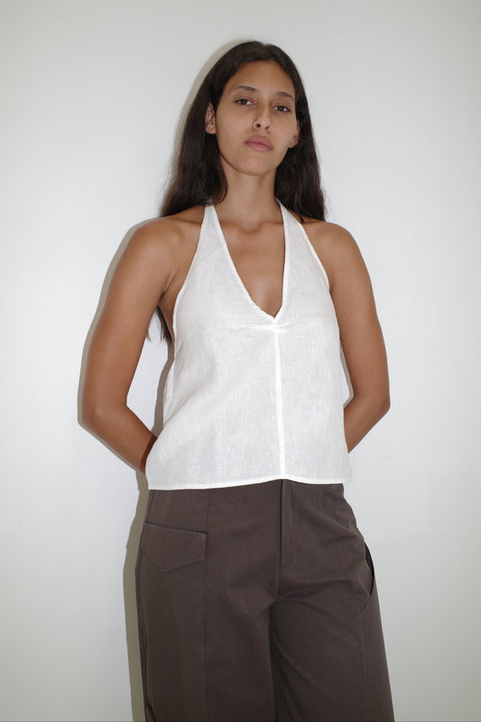 A person with long dark hair stands against a plain white wall, wearing the Linen Pinched Halter Top - White and brown pants, hands behind their back and a neutral facial expression.