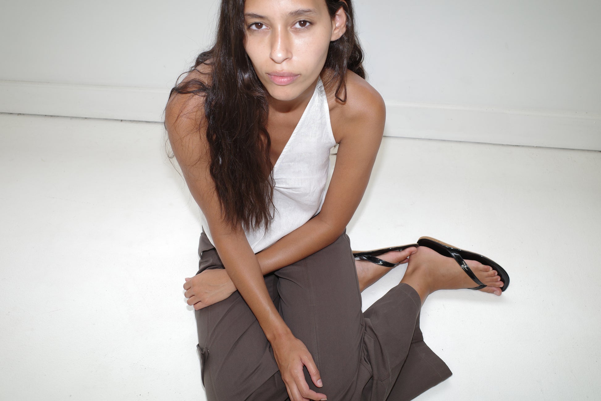 A person with long dark hair sits on the floor against a white background, wearing a Linen Pinched Halter Top - White, brown pants, and black sandals, looking up at the camera with a neutral expression.