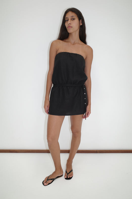 A woman with long dark hair stands against a white wall, wearing the Strapless Linen Dress - Black from Deiji Studios with a tie waist and black flip-flops, looking at the camera with a neutral expression.