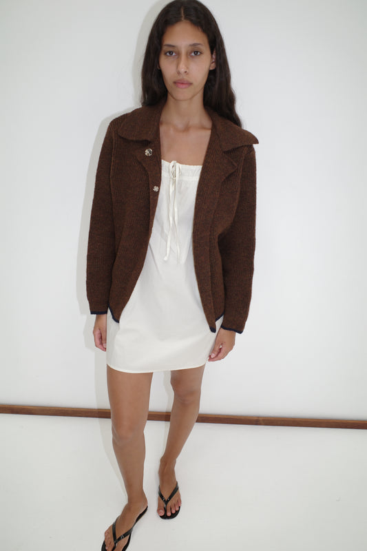A woman stands against a white wall wearing the Light Dress - White from Deiji Studios under a brown knit jacket, paired with black flip-flops. Her long wavy dark hair and neutral expression give the look an effortless elegance.