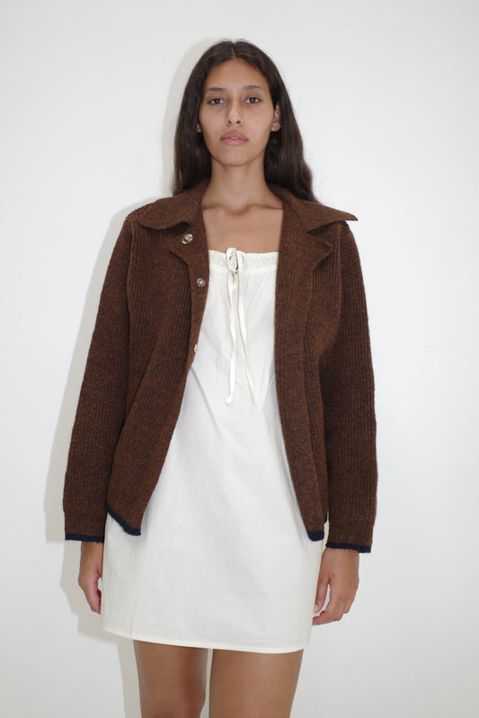 A woman with long dark hair and a neutral expression stands against a plain white wall wearing the Light Dress - White by Deiji Studios, an organic cotton mini dress with a front tie, layered under a brown knit jacket.