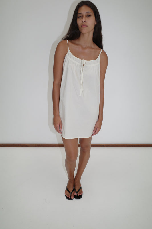 A woman with long dark hair wears the Light Dress - White, a simple sleeveless mini dress with thin straps by Deiji Studios, paired with black sandals, standing against a plain white background.