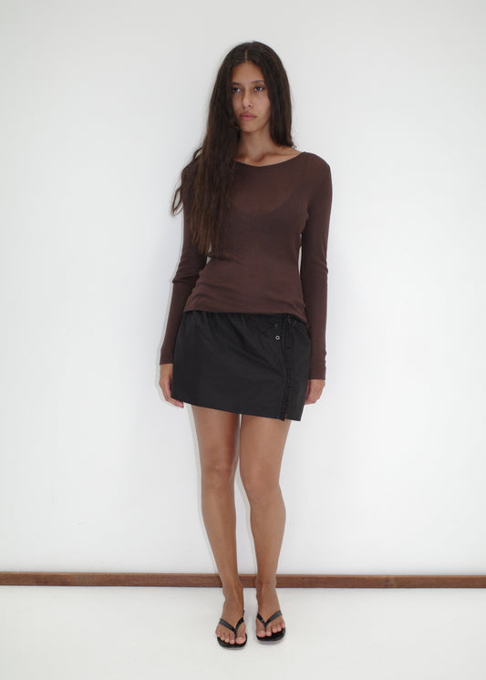 A woman with long brown hair stands against a white wall, wearing a Knitted Top - Dark Brown, a black mini skirt, and black flip-flops. She faces forward with a neutral expression.