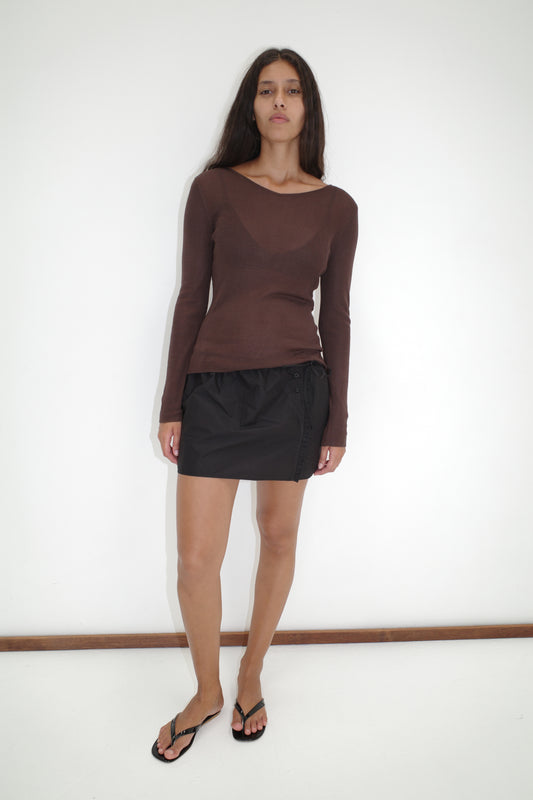 A woman with long dark hair stands against a white wall, wearing the Knitted Top - Dark Brown from Deiji Studios, a black mini skirt, and black flip-flops. She gazes at the camera with a neutral expression.