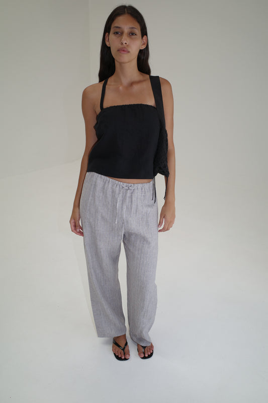 A woman with long dark hair wears a black sleeveless top, Double Waist Linen Pant in Haze Blue, and black sandals, standing in a minimalist white room and looking at the camera with a neutral expression.
