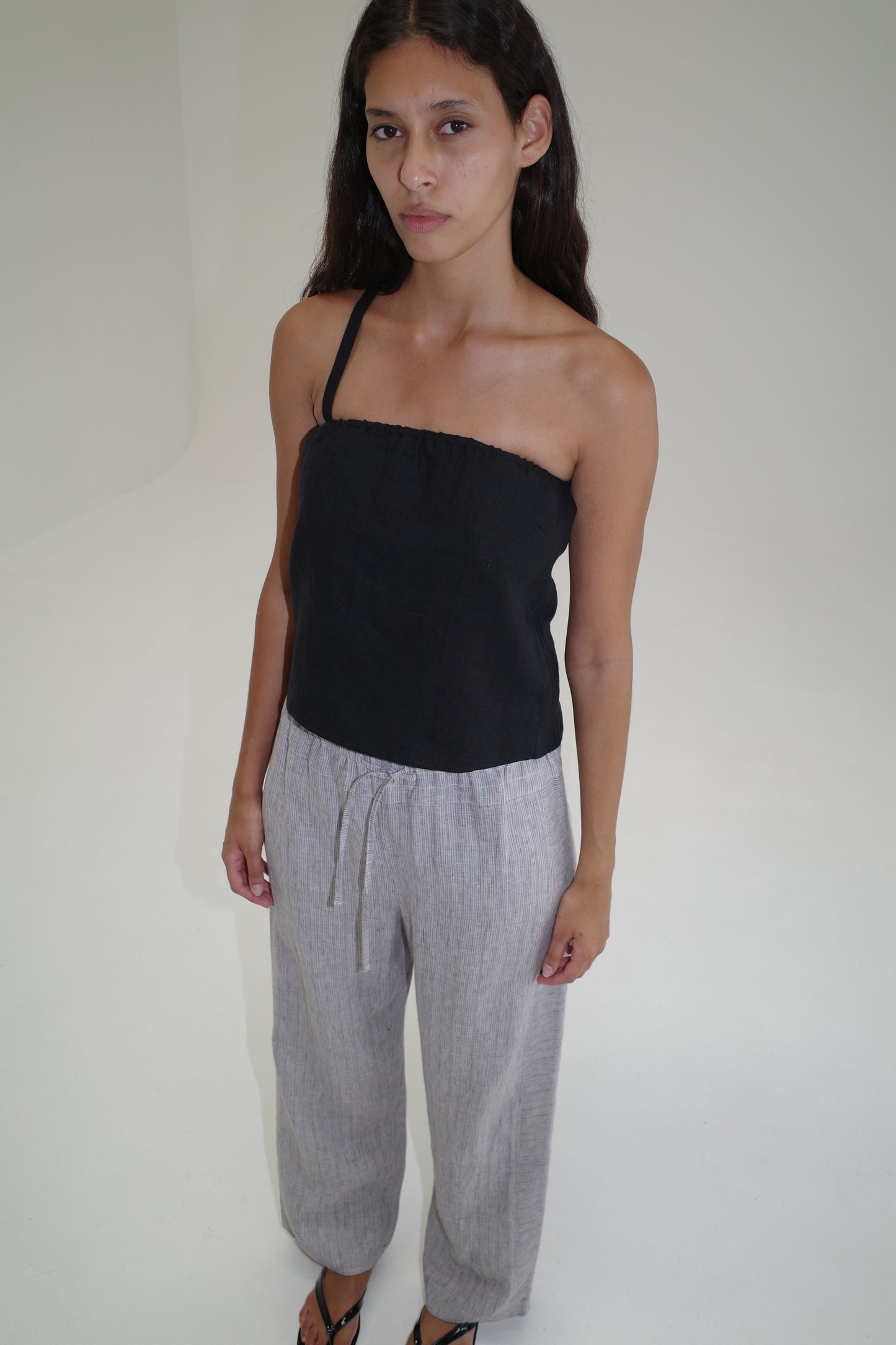 A woman with long dark hair stands in a minimalist, light-colored room, wearing a black strapless top and the Double Waist Linen Pant in Haze Blue, crafted from organic French linen. She gazes slightly away from the camera with a neutral expression.