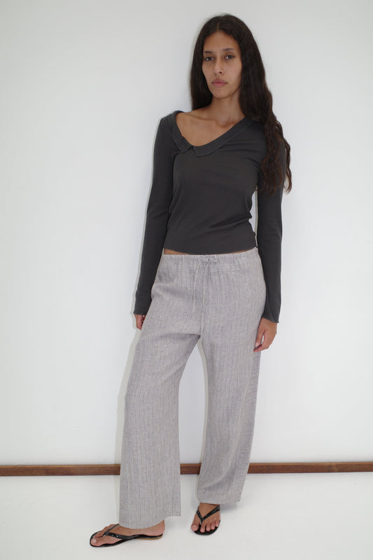A woman with long dark hair stands against a white wall, wearing a dark asymmetrical long-sleeve top and Double Waist Linen Pant in Haze Blue. She finishes the look with black flip-flops and a neutral expression.