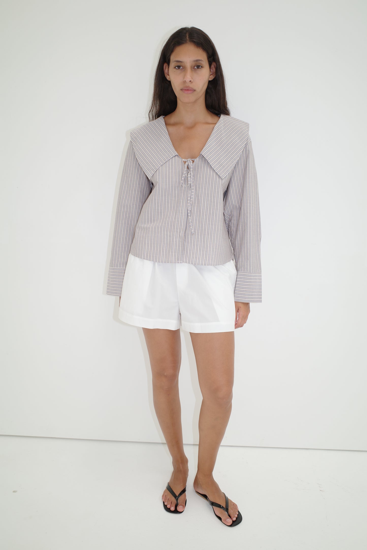 A woman with long dark hair and a neutral expression stands against a white wall, wearing the Collared Tie Shirt - Seas Stripe, featuring an oversized striped design with a large sailor collar, paired with white shorts and black flip-flops.