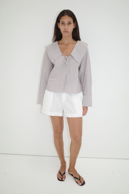 A woman with long dark hair and a neutral expression stands against a white wall, wearing the Collared Tie Shirt - Seas Stripe, featuring an oversized striped design with a large sailor collar, paired with white shorts and black flip-flops.