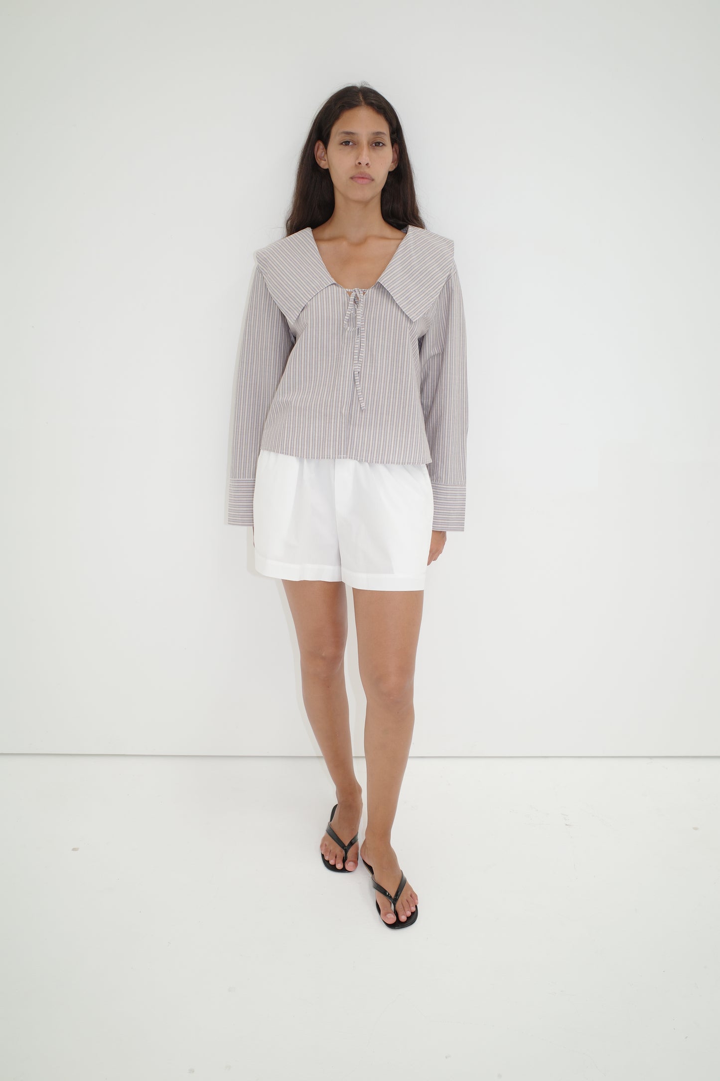 A woman with long dark hair and a neutral expression stands against a white wall, wearing the Collared Tie Shirt - Seas Stripe—an oversized shirt with bold stripes and a large collar—paired with white shorts and black flip-flops.