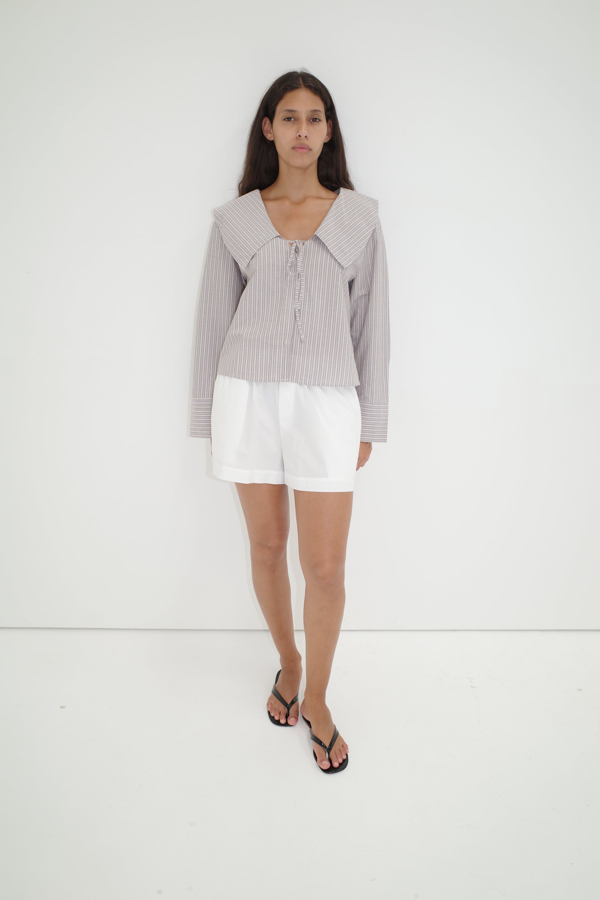 A woman with long dark hair and a neutral expression stands against a white wall, wearing the Collared Tie Shirt - Seas Stripe—an oversized shirt with bold stripes and a large collar—paired with white shorts and black flip-flops.