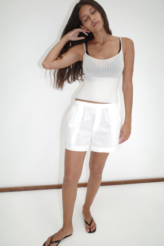 A woman with long brown hair stands against a white wall, eyes closed and hand in her hair, wearing an Organic Cotton Boxer - White from Deiji Studios paired with a sleeveless top and black bra.