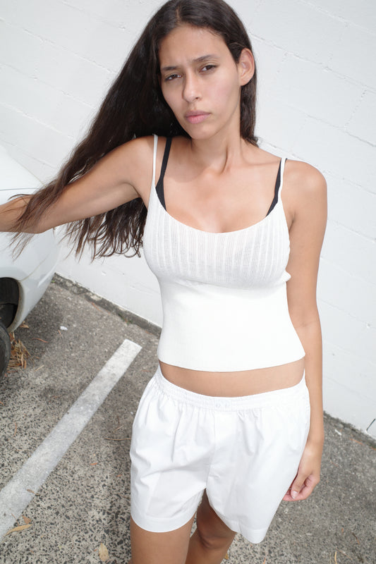 A woman with long brown hair stands outdoors on pavement, wearing a white sleeveless top over a black bra and the Organic Cotton Boxer - White. She looks at the camera with a neutral expression, against a white brick wall.