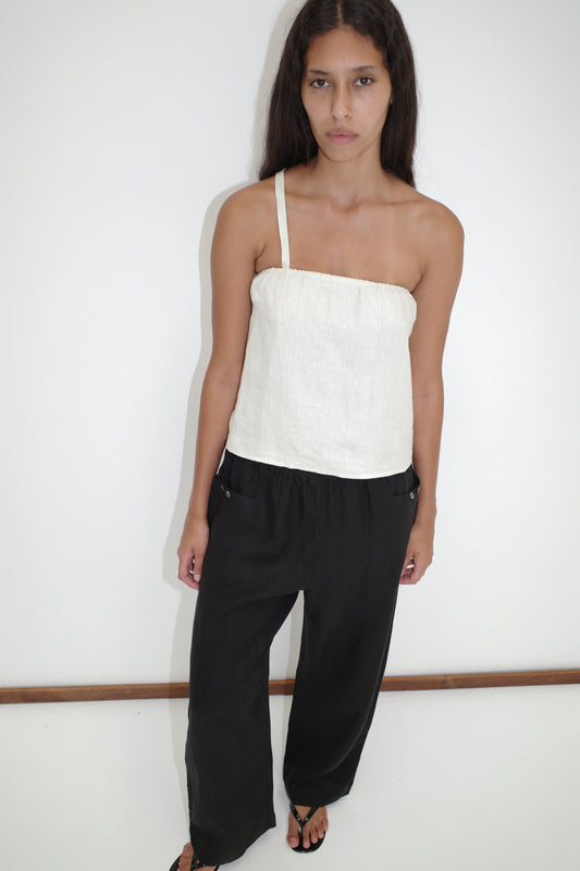 A woman with long dark hair stands against a white wall, wearing the Button Strap Top in Pastel Pistachio by Deiji Studios, paired with loose black pants and black sandals. She has a neutral expression and looks slightly up at the camera.