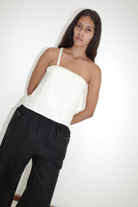 A woman with long dark hair stands against a white wall, wearing the Button Strap Top - White by Deiji Studios and loose black pants. She gazes calmly at the camera with her arms behind her back.