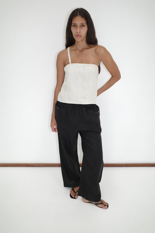 A woman stands against a white wall wearing the Button Strap Top - Pastel Pistachio by Deiji Studios, paired with loose black pants and black sandals. She has long dark hair, a neutral expression, and one hand in her pocket.