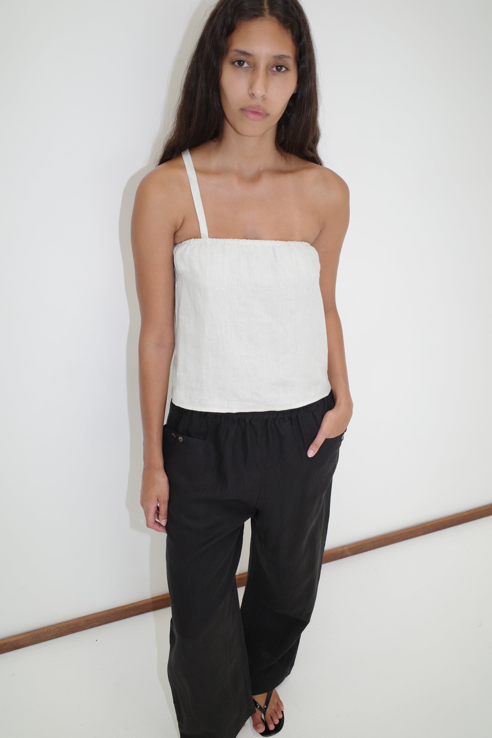 A woman with long dark hair stands against a white wall, wearing Deiji Studios’ Button Strap Top in white, loose black pants, and black sandals, with her hands in her pockets.