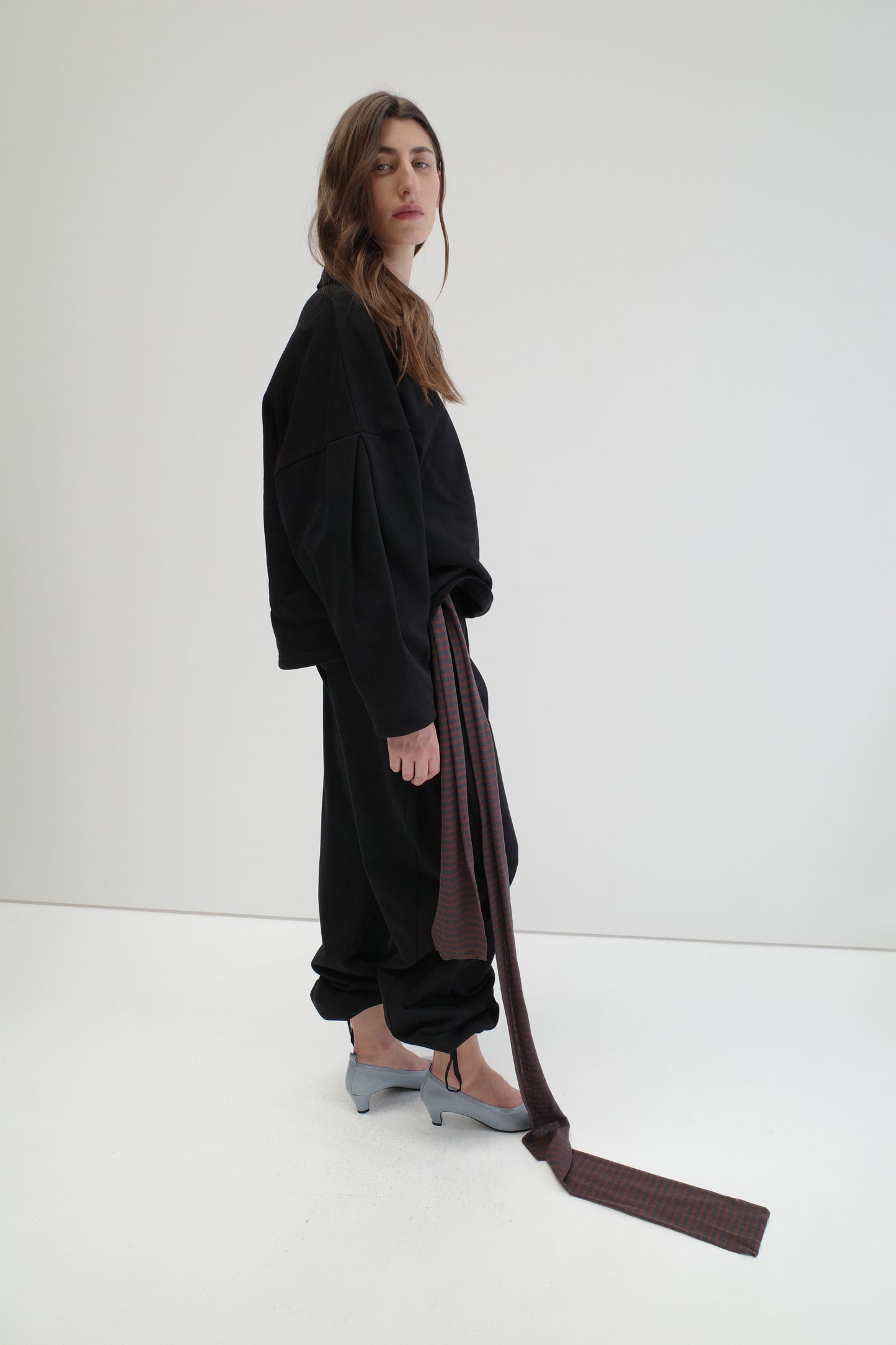 A woman in a minimalist white space wears loose black clothing, slouchy pants, a long checked brown belt, light blue heels, and the Knit Scarf - Brown Grey Stripe draped around her neck. She has long brown hair and a neutral expression.