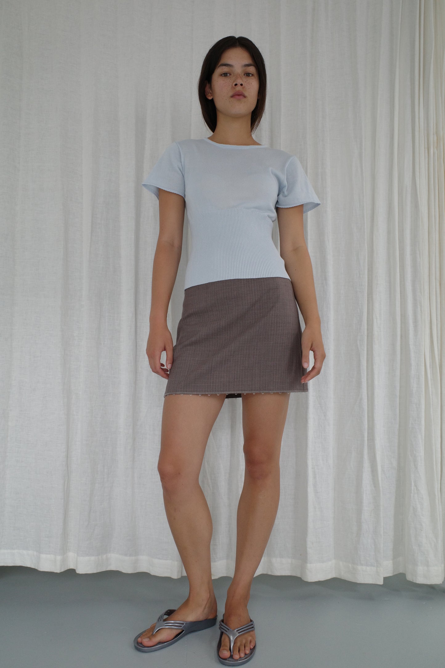 A woman stands before white curtains, confidently facing the camera in Deiji Studios’ Trim Knit Tee - Ice Blue, paired with a brown mini skirt and blue sandals.
