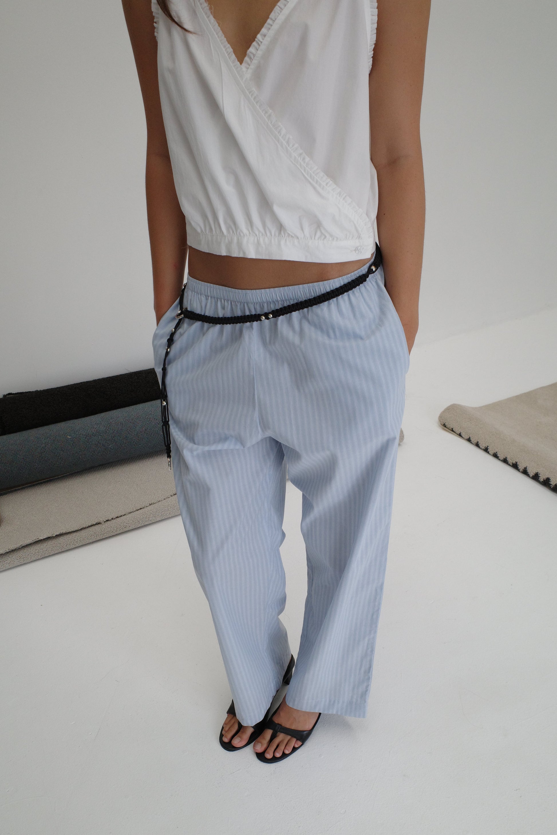 A person stands indoors wearing black sandals, a sleeveless white wrap top, and the Ease Trouser in light blue pinstripes with hands in their pockets; their face is not shown.