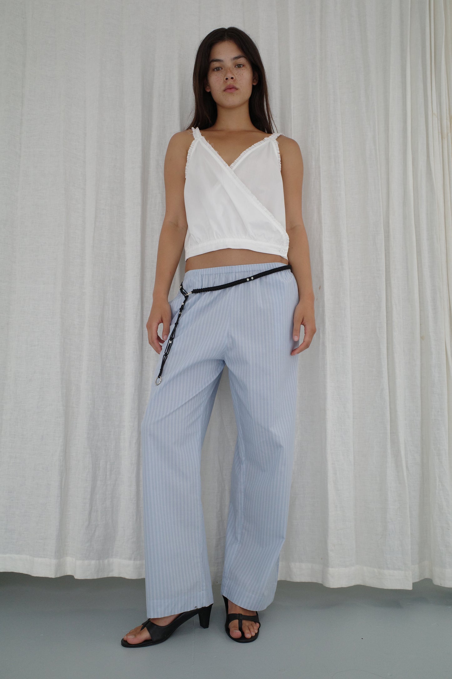 A person stands before white curtains wearing a Deiji Studios white sleeveless wrap top, Ease Trouser - Lake Blue Stripe with a black belt, and black heeled sandals. They have straight dark hair and a neutral expression.