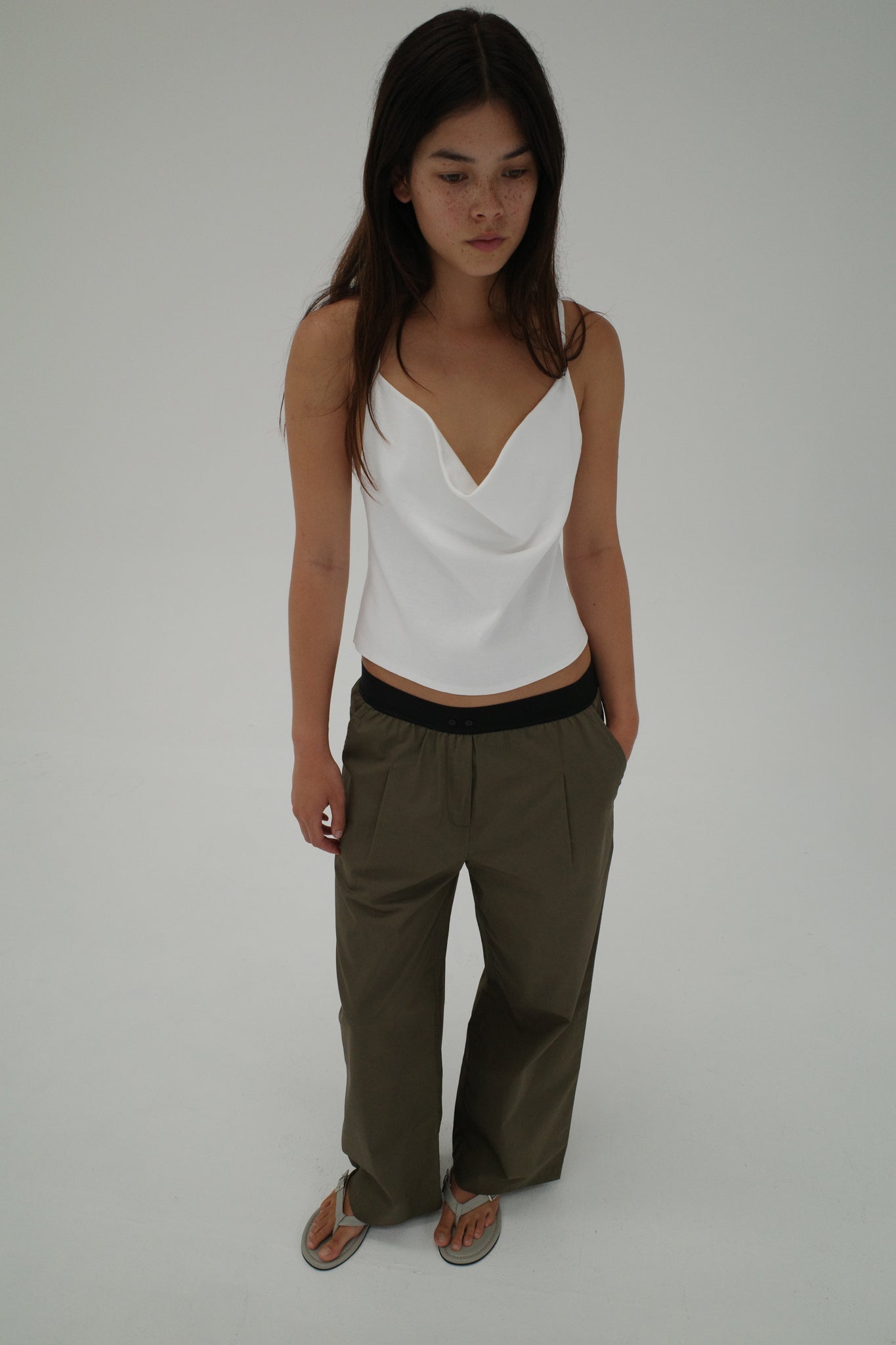 A woman with long dark hair wears a white sleeveless top, Deiji Studios' Contrast Waist Pant - Olive, and sandals as she stands against a plain white background.