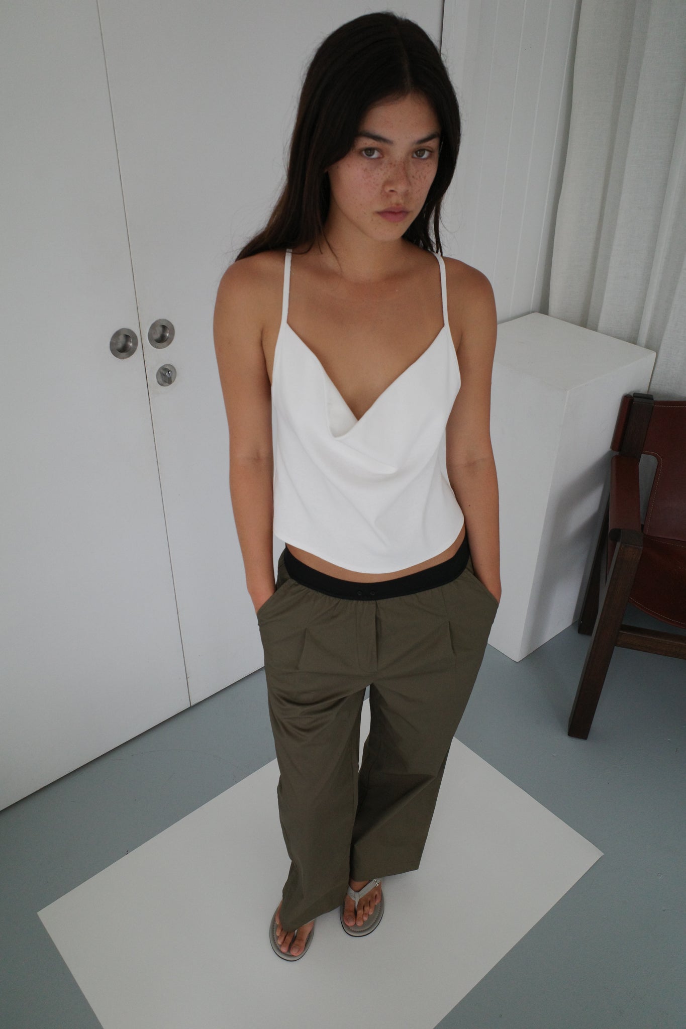 A woman with long dark hair wears a white camisole and Contrast Waist Pant, standing with hands in her pockets on a white mat in a minimal room with white walls and simple furniture.