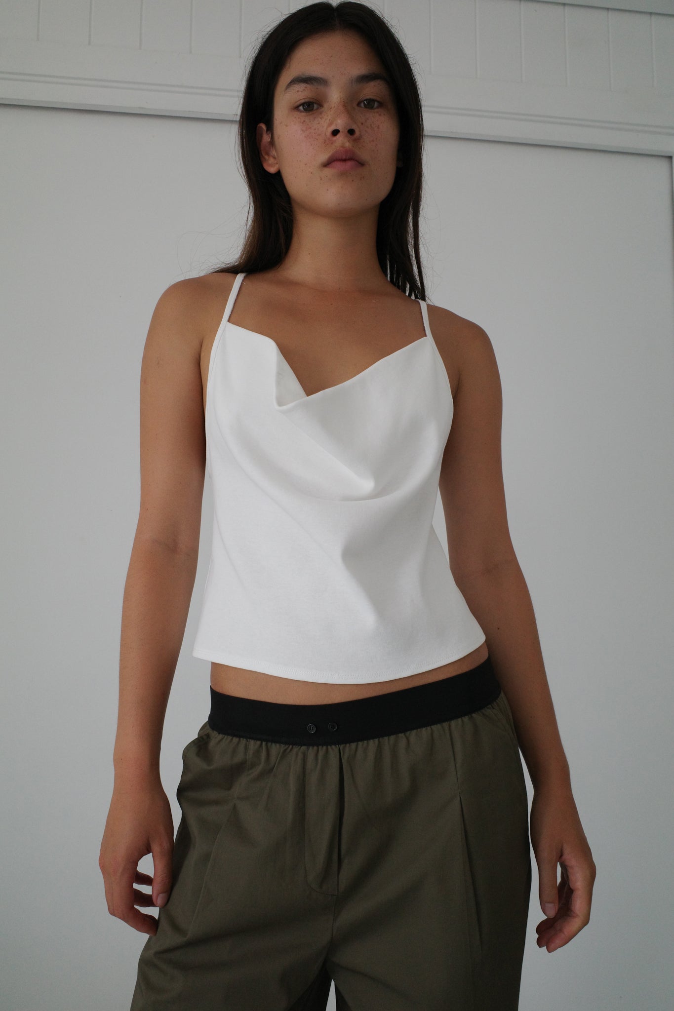A woman wears a white draped camisole and the Contrast Waist Pant - Olive, standing confidently before a plain white background.