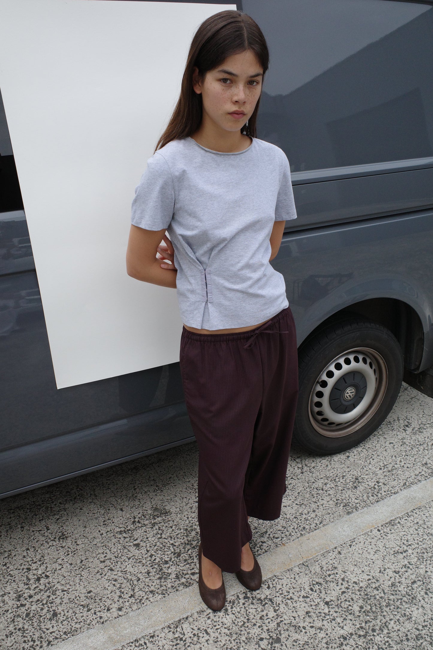 A woman with long brown hair stands in front of a gray van, wearing the Deiji Studios Pinch Tee in grey, loose dark brown pants, and brown shoes. Her hands are behind her back as she looks slightly to the side.