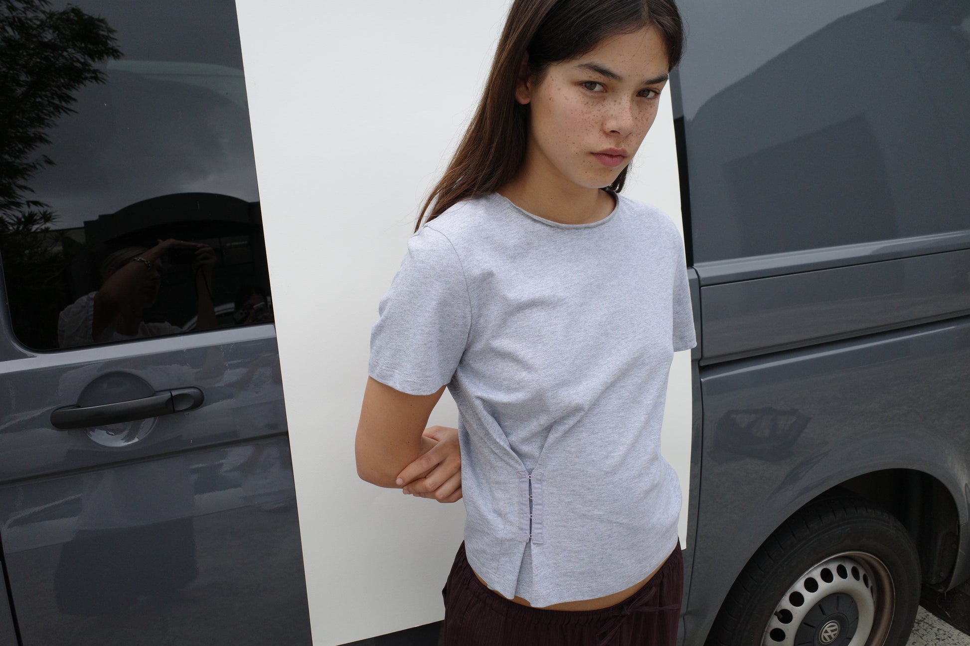 A woman with long dark hair wears the Pinch Tee - Grey and dark pants, standing outdoors in front of a gray van and looking at the camera with a neutral expression.
