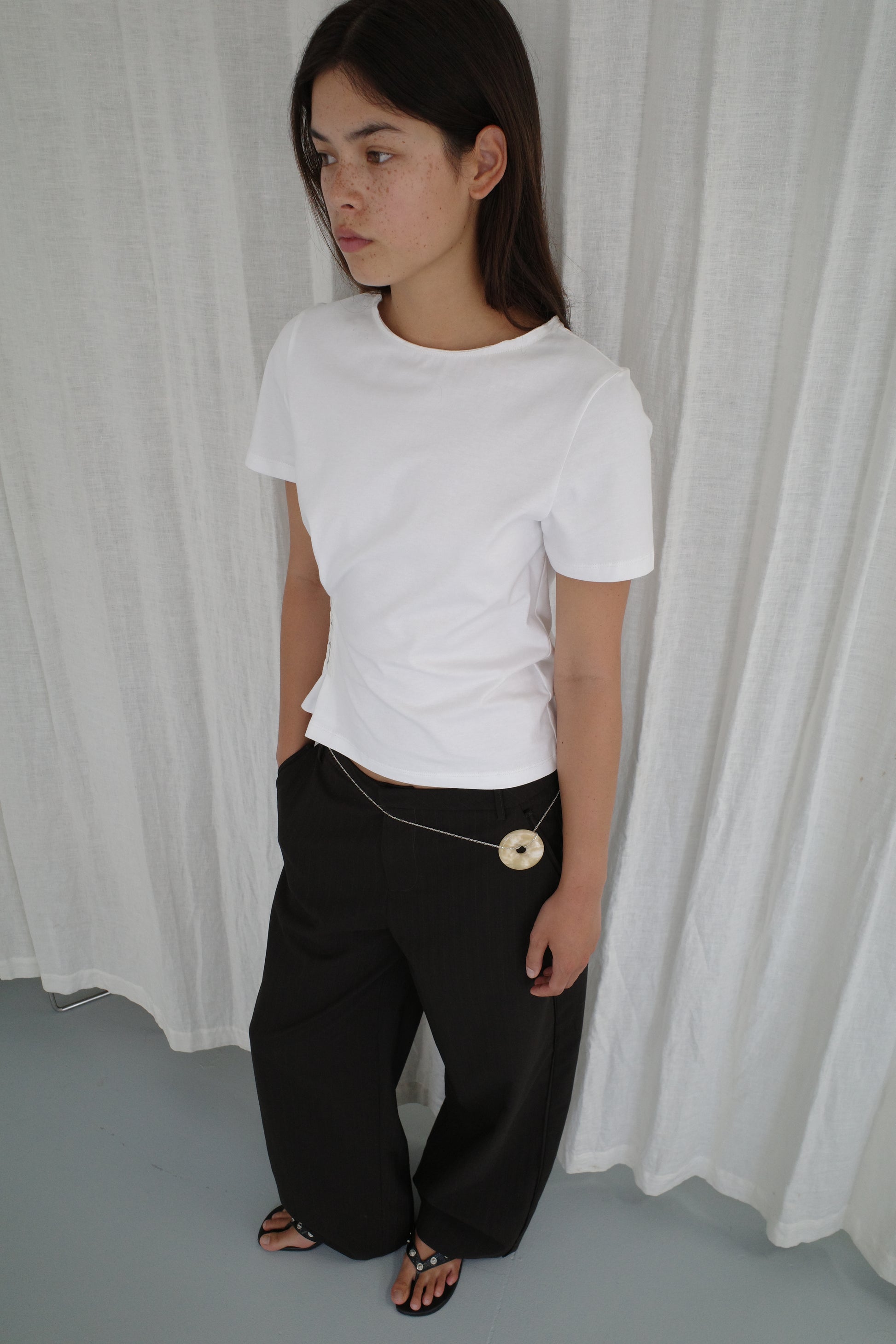 Model wearing the Pinch Tee - White from Deiji Studios, paired with wide-leg black trousers with a chain belt and round disc pendant, plus black strappy sandals, standing against a white linen curtain backdrop.