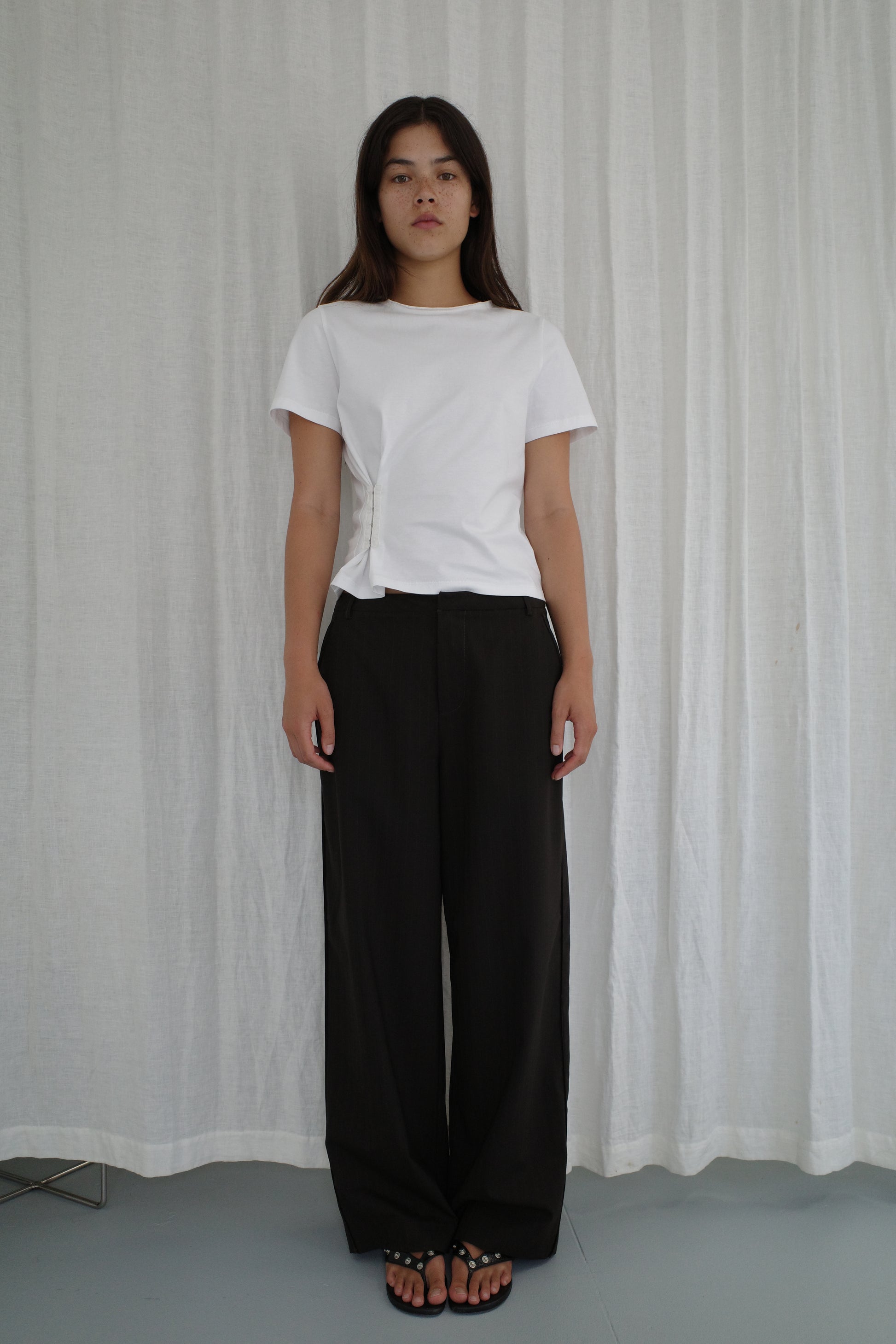 A woman with long dark hair stands in front of white curtains, wearing the Pinch Tee - White by Deiji Studios, loose black pants, and black sandals. She has a neutral expression and her arms are relaxed at her sides.
