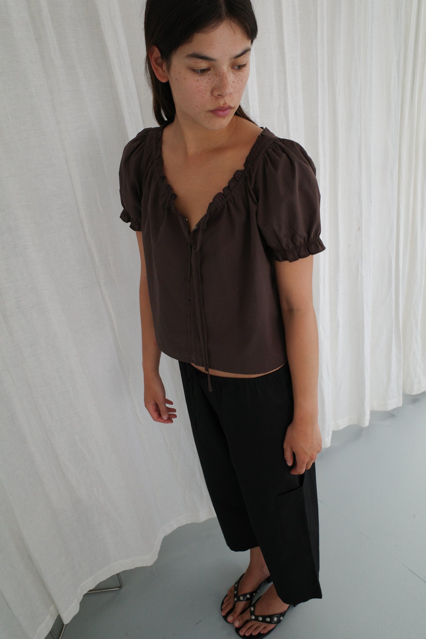 A person stands indoors on a light floor, wearing the Close Shirt - Sepia by Deiji Studios, black wide-leg pants, and black flip-flops. They look down and away from the camera, with a white curtain backdrop.