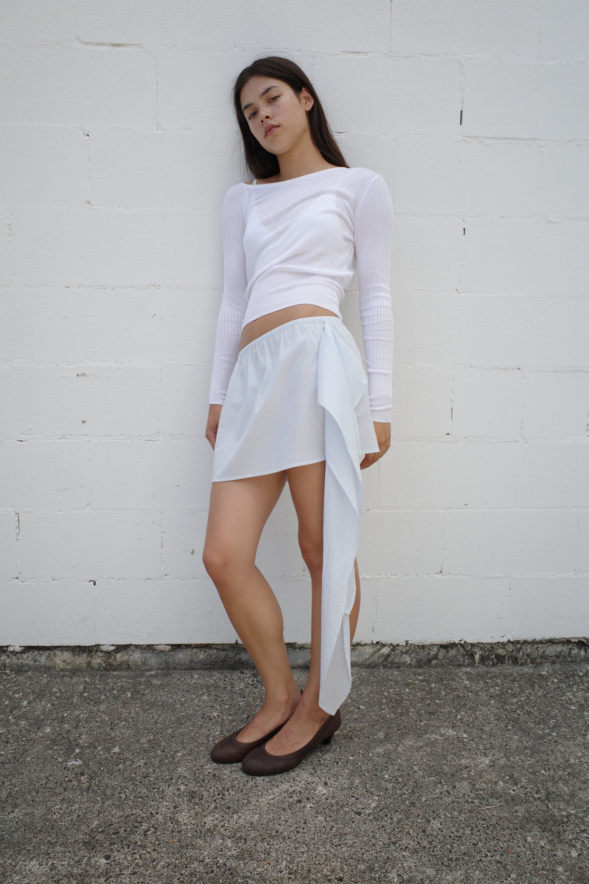 A woman with long dark hair and a neutral expression stands against a white brick wall, wearing the Deiji Studios Shoulder Knit Top in white, an asymmetrical draped white skirt, and brown flat shoes.