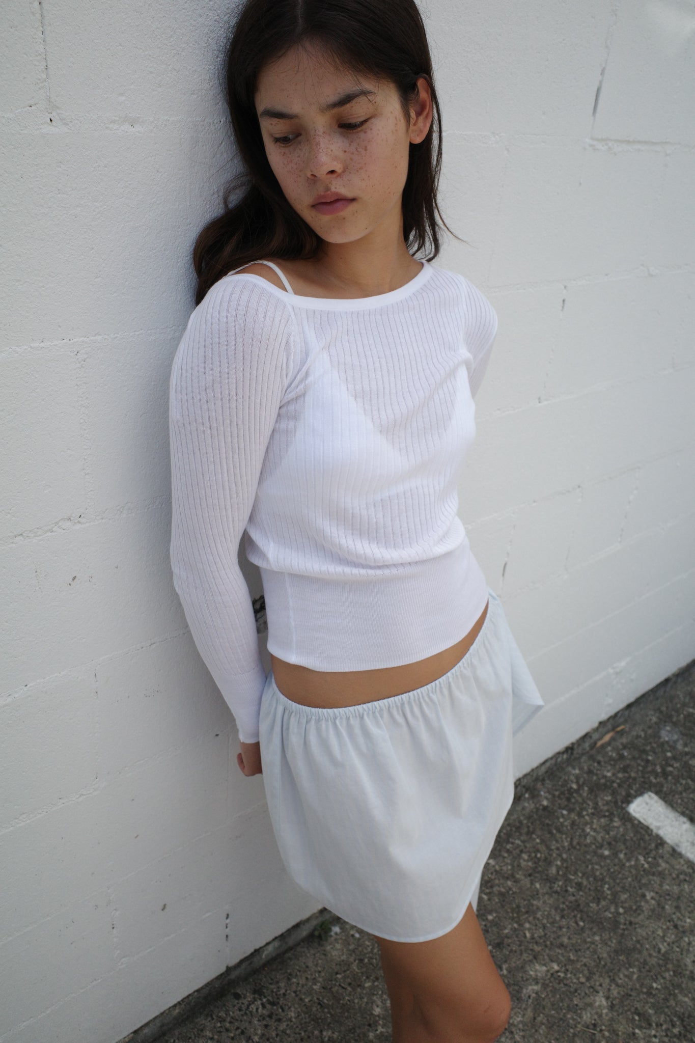 A woman with long dark hair stands against a white textured wall, wearing the Shoulder Knit Top - White and a light, high-waisted skirt. She gazes downward with a pensive expression.